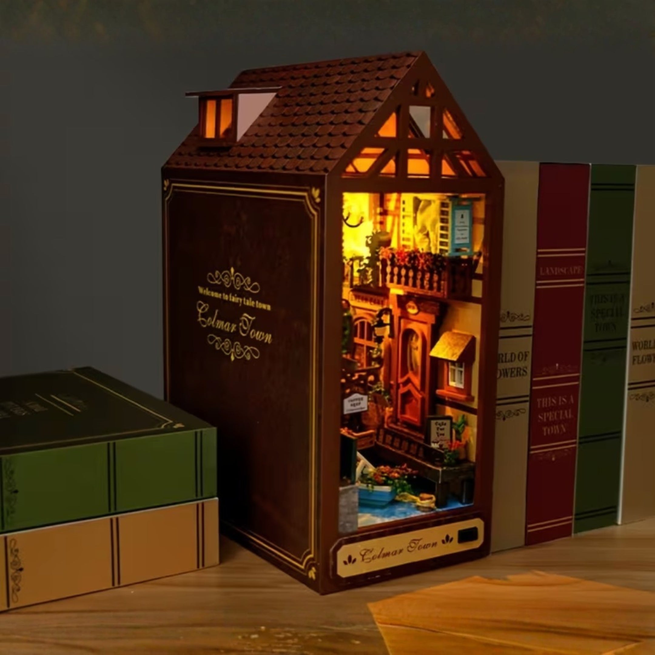 Миниатюрен book nook модел на къща с LED осветление, поставен между книги на рафт, фентъзи сцена с малко градче и детайлна архитектура