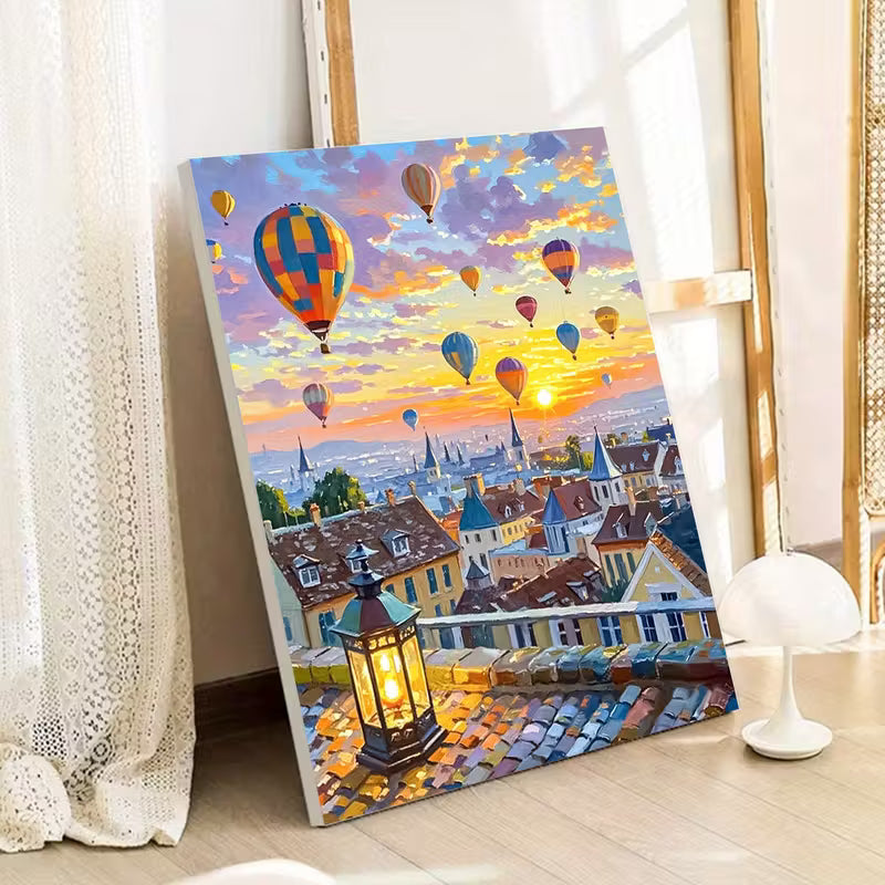 Рисуване по номера с градски пейзаж при залез, платното е облегнато до стена до прозорец с лека завеса, изобразени са покриви на къщи в топли цветове, светещ фенер на преден план и множество цветни балони с горещ въздух, издигащи се над града под небе в златисти, розови и сини нюанси, създаващи спокойна и романтична атмосфера.