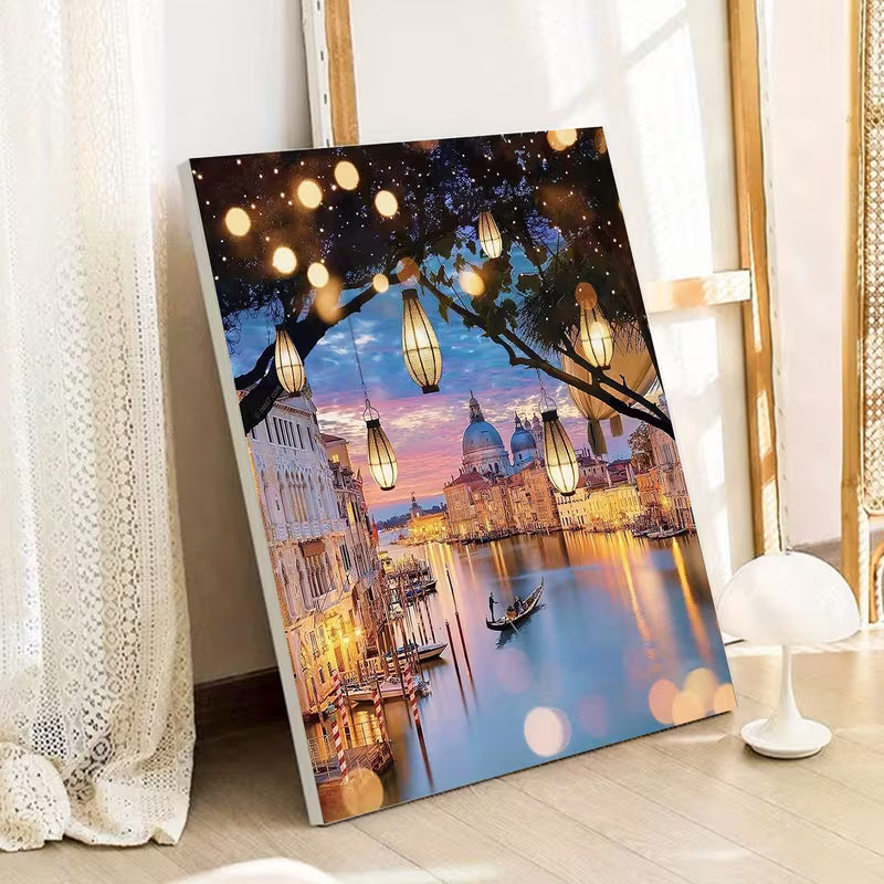 Рисуване по номера с вечерен пейзаж от Венеция, изобразяващ канал с гондола, исторически сгради и куполи, осветени от топли висящи лампи сред клоните на дърво, с отражения във водата и романтична атмосфера, показано като готово платно, облегнато до стена в интериор.