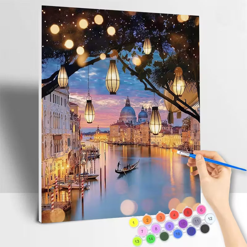 Рисуване по номера с романтична вечерна сцена от Венеция, канал с гондола, исторически сгради и куполи на заден план, осветени от топли лампи, висящи сред клоните на дърво, отражения във водата и меко небе в синьо и розово, показано в процес на рисуване с четка и номерирани бои.