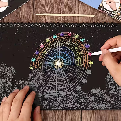 Скреч картина в процес на работа, показваща виенско колело с цветни светлини върху черен фон, докато ръка изтрива покритието със скреч писалка и разкрива ярките детайли на нощната сцена.