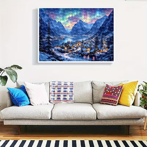 Пъзел с зимен планински пейзаж, подреден в рамка на стената над диван, със заснежено селце край езеро и планини под цветно нощно небе.