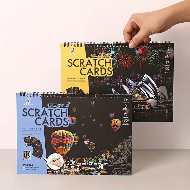 Два комплекта скреч картини с 10 изображения всеки, показани отпред, с нощни градски сцени, фойерверки и балони с горещ въздух, спирално подвързани, държани с ръка за сравнение на дизайните и размера.