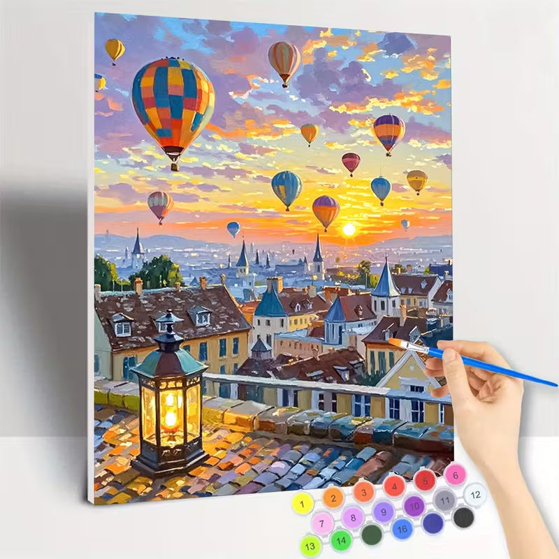 Рисуване по номера с романтичен градски пейзаж, залез над покриви на къщи и катедрали, множество цветни балони с горещ въздух в небето, светещ уличен фенер на преден план и ръка, която оцветява платното с четка и номерирани бои.
